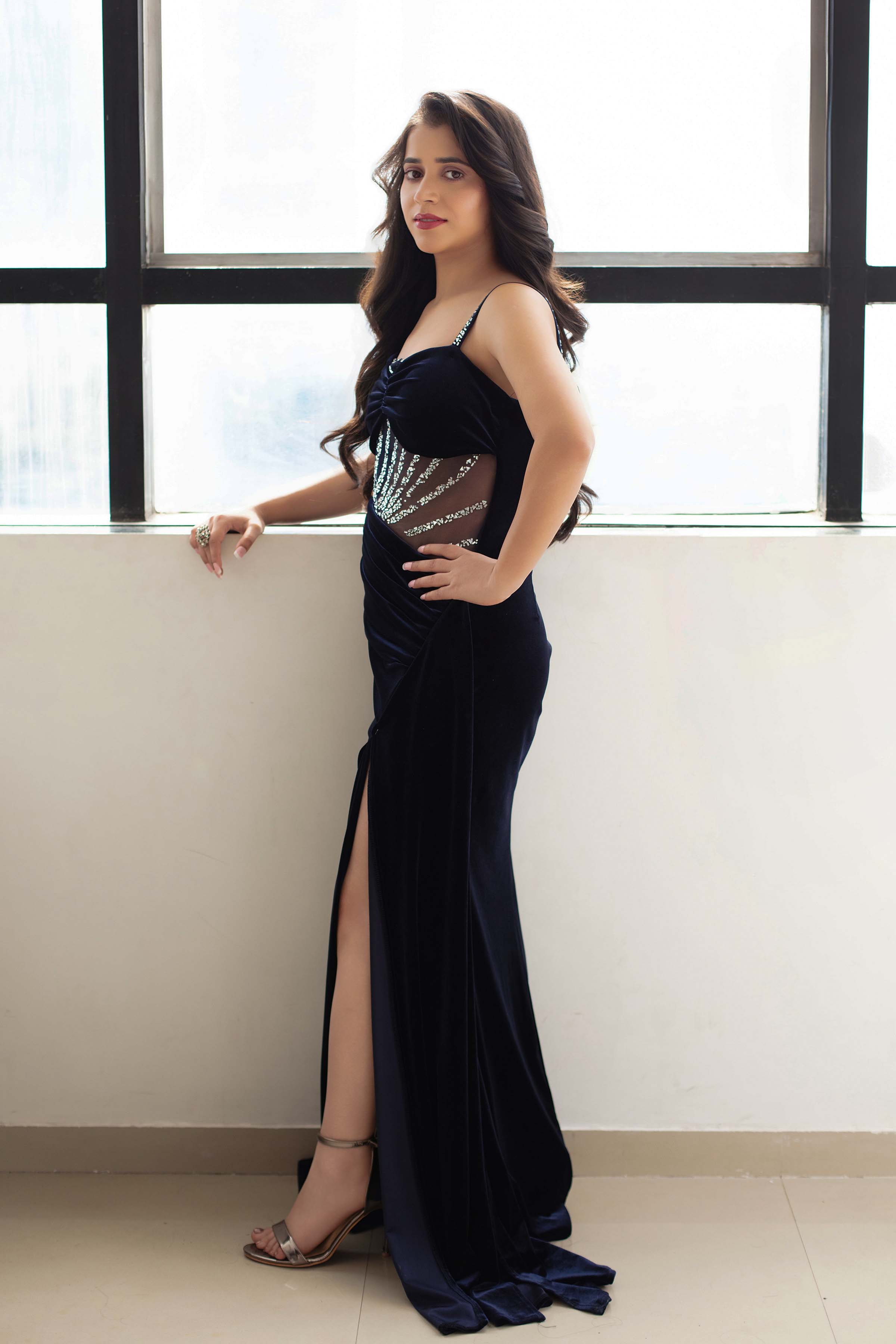 Neeru Sharma in a navy velvet gown with a thigh-high slit, by a window